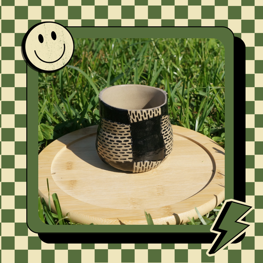A handmade ceramic cup with a checkered inlay design, displayed on a wooden stand with a cheerful face and lightning bolt graphics around it.