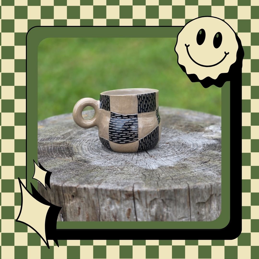 Ceramic cup with black patterns on a wooden surface, framed by a green and white checkered border with a smiley face.