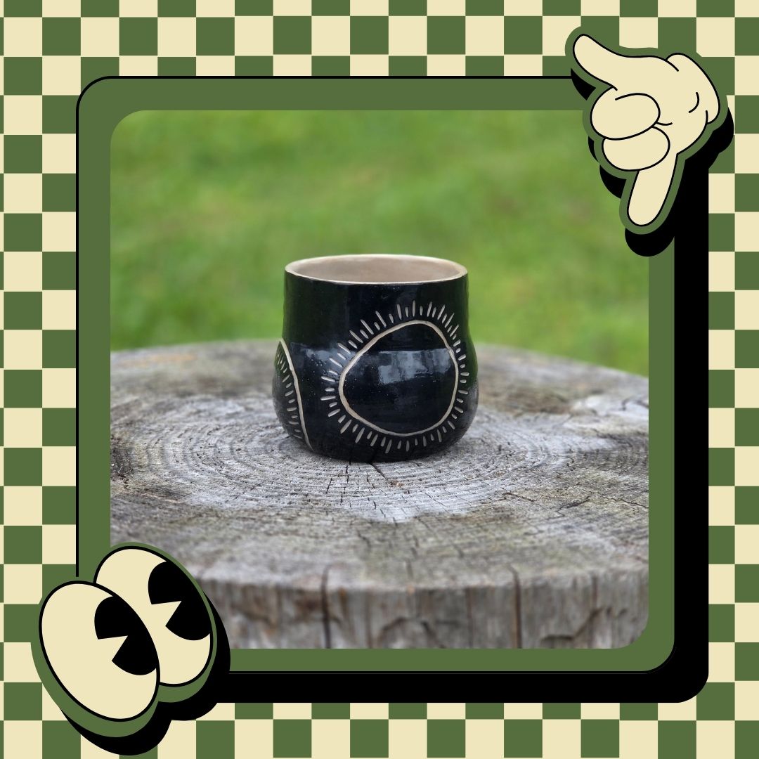 Black ceramic mug with sun design on a wooden stump outdoors, framed by a green and white checkered border.