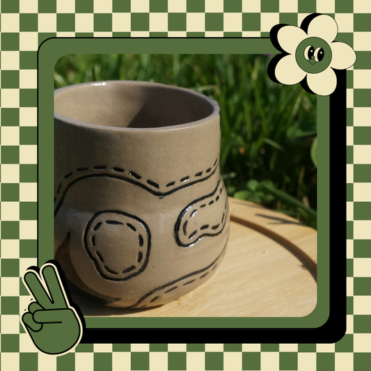 A beige ceramic mug with black abstract inlay designs, displayed on a wooden round tray, with a peace sign and a cartoonish face illustration in the corner.
